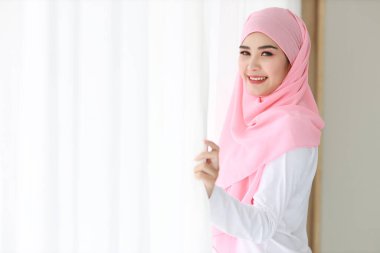 Yan görünüşlü güzel Asyalı Müslüman kadın, beyaz uyku giysisi giyiyor, sabahleyin kalktıktan sonra pencerenin önünde dikiliyor. Pembe tesettürlü tatlı genç kız kameraya bakarken rahatlıyor.