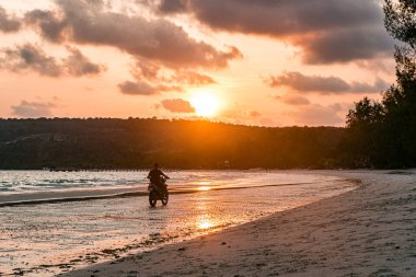 Tropik bir adada gün batımında motosiklet süren bir adam.