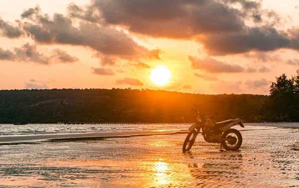 Gümüş bir motosiklet gün batımında sahilde duruyor.