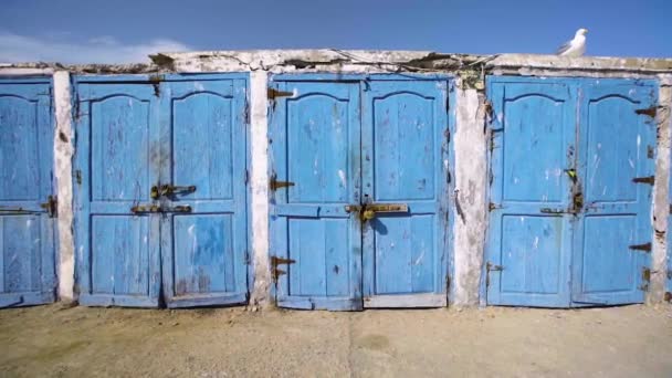 Suivi à la main le long d'une rangée de portes bleues peintes vers des hangars de stockage à Essaouira, Maroc — Video