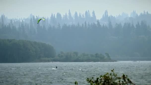 Eine Landschaftsaufnahme von Kiefern mit einem großen See im Vordergrund und einer Person beim Kitesurfen — Stockvideo
