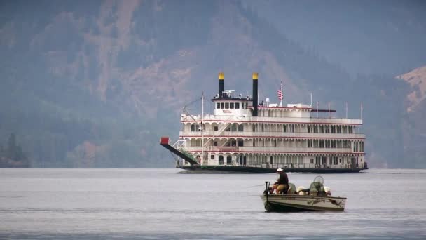 A man fishing on a small boat, with a ferry passing behind — Stockvideo