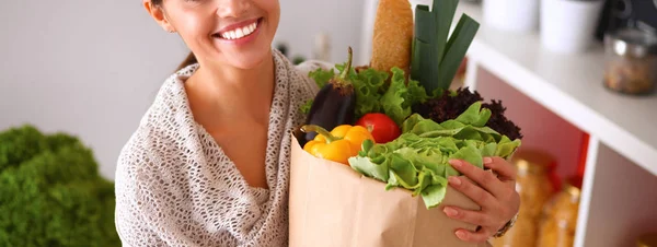 Mujer joven sosteniendo bolsa de la compra de comestibles con verduras de pie en la cocina —  Fotos de Stock