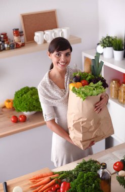 Sebzeli alışveriş poşeti taşıyan genç bir kadın mutfakta duruyor.