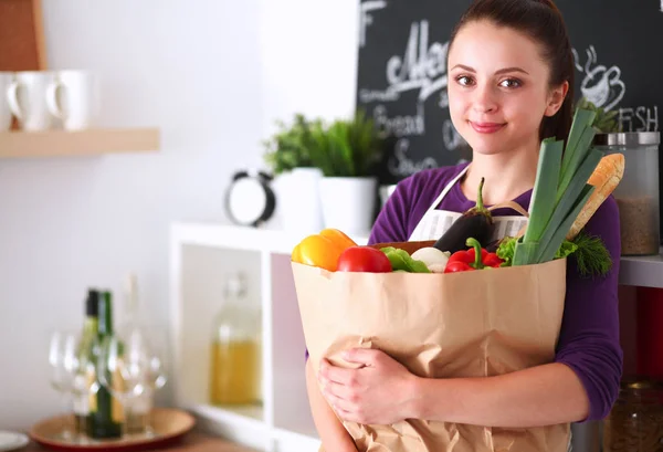 Mujer joven sosteniendo bolsa de la compra de comestibles con verduras de pie en la cocina —  Fotos de Stock