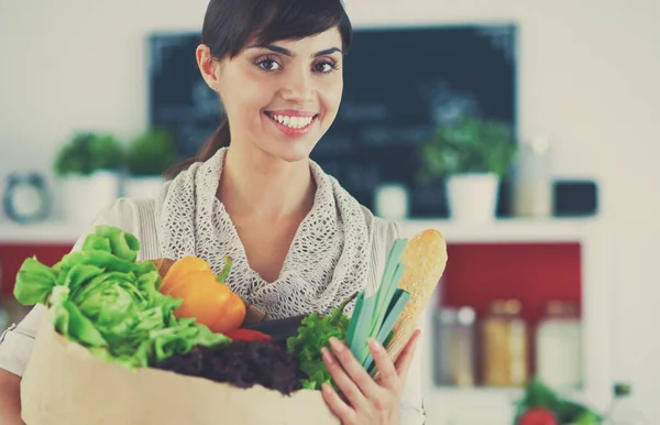 Mujer joven sosteniendo bolsa de la compra de comestibles con verduras de pie en la cocina — Foto de Stock