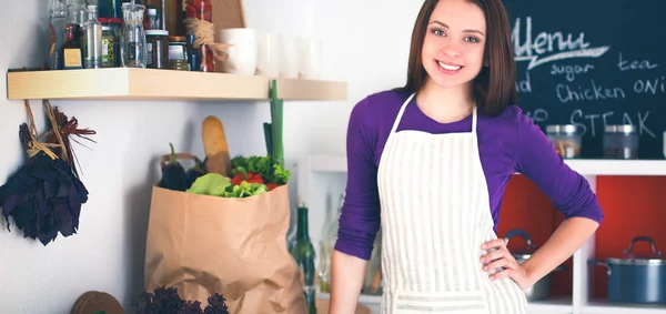 Mladá žena stojící ve své kuchyni s pytel s potravinami v pozadí. — Stock fotografie