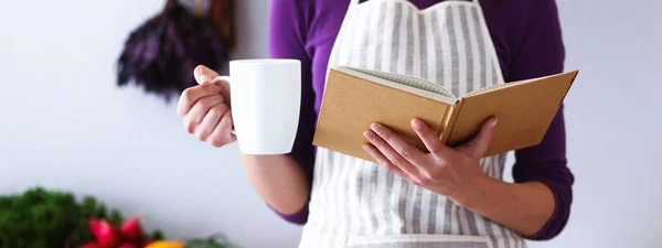 Una giovane donna in piedi nella sua cucina a bere tè e tenere un libro di cucina. — Foto Stock