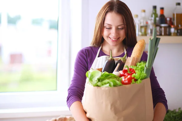 Mujer joven sosteniendo bolsa de la compra de comestibles con verduras de pie en la cocina — Foto de Stock