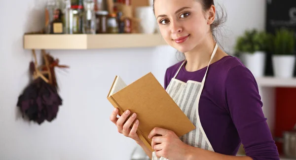 Uma jovem mulher de pé na cozinha lendo um livro de receitas. — Fotografia de Stock