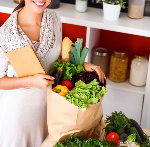 Mujer joven sosteniendo bolsa de la compra de comestibles con verduras de pie en la cocina —  Fotos de Stock