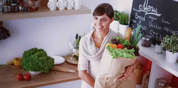 Jovem segurando supermercado saco de compras com legumes de pé na cozinha — Fotografia de Stock
