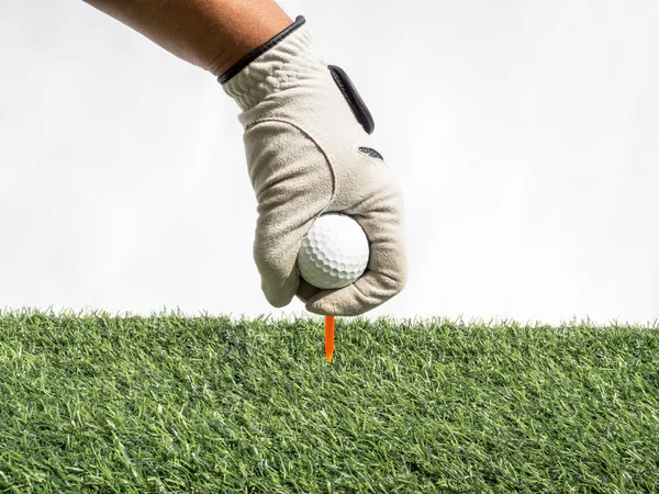 Foto de uma mão Colocar bola de golfe em um tee com grama artificial e fundo branco — Fotografia de Stock