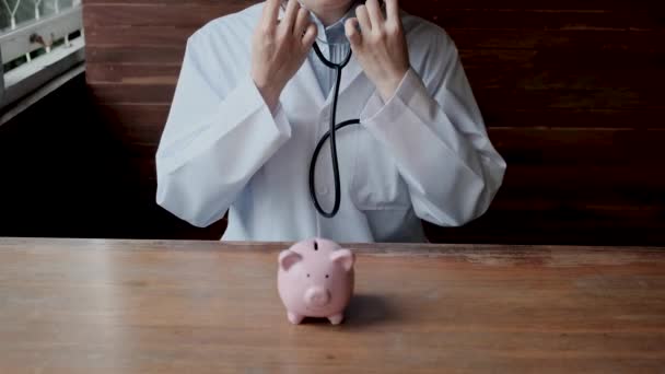医者はピンクの貯金箱をチェックするために聴診器を使用しました 将来の事業計画と健康保険の概念のためにあなたの財政の健康をチェック — ストック動画