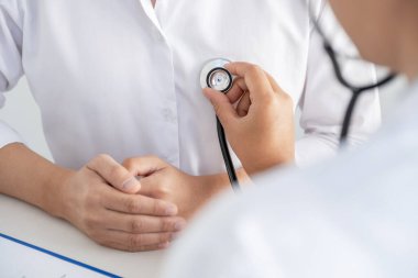 Doktor, hastayı steteskopla, tansiyon monitörüyle muayene eder ve sonuçları kaydeder..