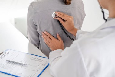 Doktor, hastayı steteskopla, tansiyon monitörüyle muayene eder ve sonuçları kaydeder..