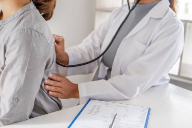 Doktor, hastayı steteskopla, tansiyon monitörüyle muayene eder ve sonuçları kaydeder..