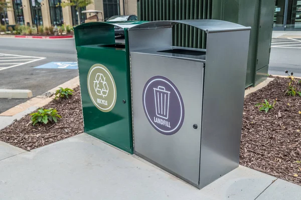 Two side by side metal garbage cans one for recyclables and the other for landfill on a cement pad in a parking lot in front of buildings angle view