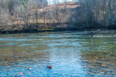 Üç yetişkin, kışın sonlarında güneşli bir günde nehirde balık tutarak eğleniyorlar.