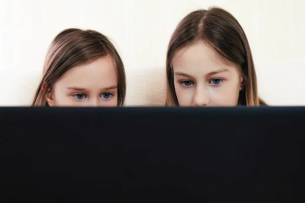 Två flickor skolflickor är noga tittar på en bärbar dator när du sitter hemma på soffan — Stockfoto
