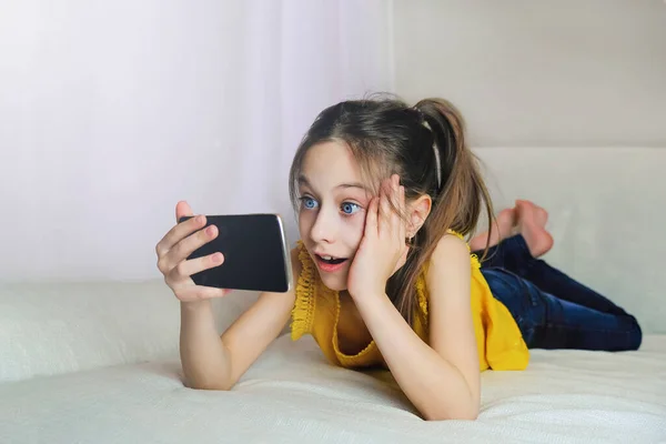 Dziewczyna otwiera usta w zaskoczeniu z przesuń na ekranie iPhone w domu leżąc na kanapie — Zdjęcie stockowe