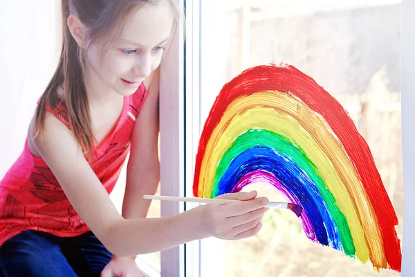 Teen girl draws a rainbow on the window a symbol of the fight against coronavirus — Stock Photo, Image