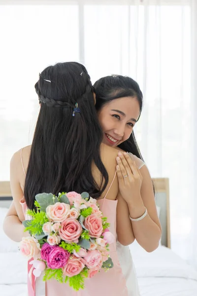 白いウェディングドレスのアジアの花嫁は笑顔でピンクのドレスで彼女の友人を包含し その後 彼女の左指にダイヤモンドリングを示し 一方で 花の美しい花束を保持します — ストック写真