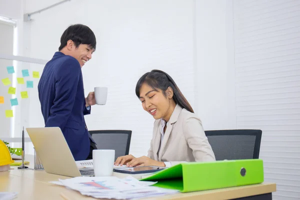 A young, professional young Asian businesswoman is sitting at her desk and sipping afternoon coffee with a male office worker working together.