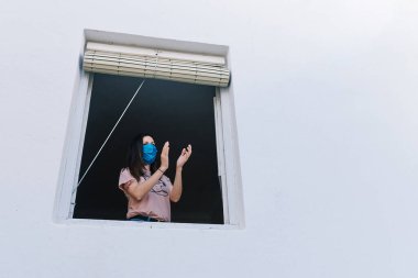 Maskeli genç esmer kadın beyaz arka planlı, evin penceresinden alkışlıyor.
