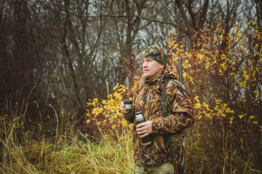 Kamuflaj kıyafetli, termoslu ve çantasında silah olan bir adam..