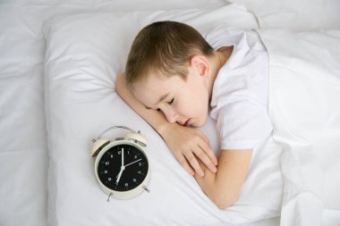 Çalar saati olan beyaz bir yatakta uyuyan bir çocuğun en iyi görüntüsü.