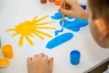 Çocuk güneşi ve gökyüzünü parlak renklerle boyar. Çocuk oturuyor.