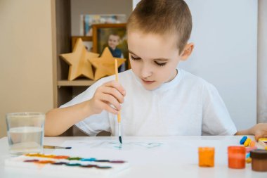Beyaz tişörtlü bir çocuk beyaz bir masada boyalarla resim çiziyor..