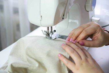 Beyaz bir masada modern dikiş makinesi. Bir kadın dikiş makinesine keten kumaş dikiyor..