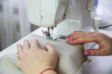 Beyaz bir masada modern dikiş makinesi. Bir kadın dikiş makinesine keten kumaş dikiyor..