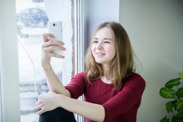 A teenage girl with long hair sits on a windowsill and looks at the screen of a mobile phone, photographs herself, communicates via video communication. It is raining outside the window.