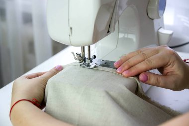 Beyaz bir masada modern dikiş makinesi. Bir kadın dikiş makinesine keten kumaş dikiyor..