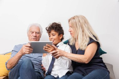 Çocuğun büyükannesi ve büyükbabasıyla kanepenin üzerinde dijital tabletle oynadığı otantik an. Evde tabletle gülümseyen aile