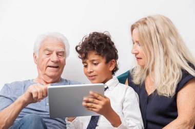 Çocuğun büyükannesi ve büyükbabasıyla kanepenin üzerinde dijital tabletle oynadığı otantik an. Evde tabletle gülümseyen aile