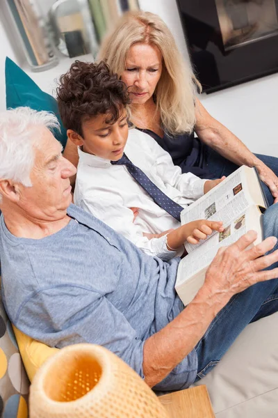 Loving grandpa and grandma with grandson reading together a dictionnary on sofa. Boy reading a book with grandparents