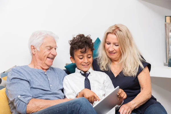 Çocuğun büyükannesi ve büyükbabasıyla kanepenin üzerinde dijital tabletle oynadığı otantik an. Evde tabletle gülümseyen aile