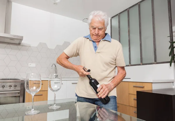 Senior man opening red wine bottle of wine with a corkscrew in a kitchen.