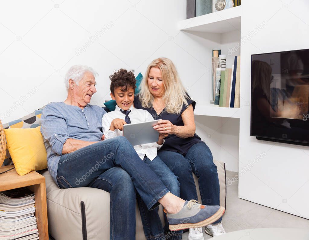 Authentic moment when boy with her grandmother and his grandfather play with a digital tablet on sofa in living room. Smiling family with tablet at home