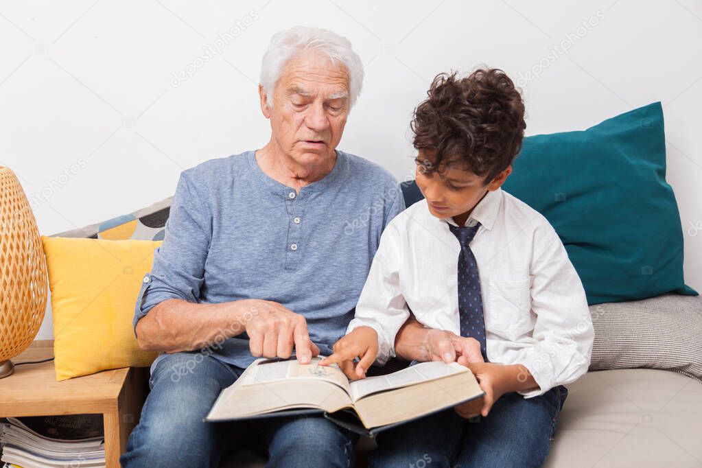 Happy grandfather with grandson reading together a book, a dictionnary on sofa. Family, cultur and joy concept