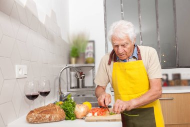 Evdeki bir mutfakta sağlıklı yiyeceklerle yemek pişiren mutlu kıdemli bir adam. Sarı masalı bir adam sebze kesiyor.