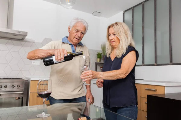 Senior man serving red wine in a glass at his wife, a beautiful blonde woman.