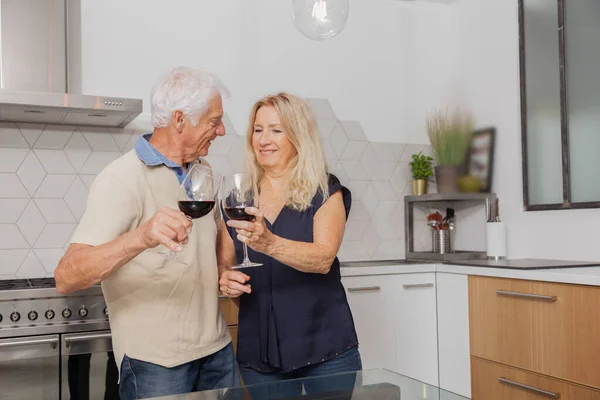Happy senior couple relax talking and drinking red wine glasses together in the kitchen at home