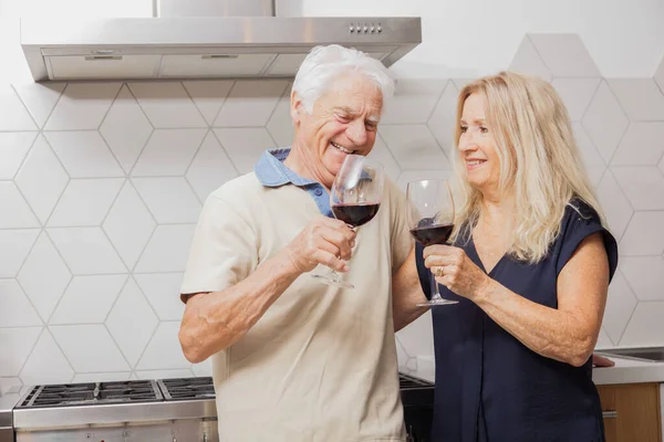 Happy senior couple relax talking and drinking red wine glasses together in the kitchen at home