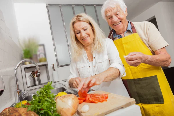 Happy senior couple cooking together with healthy food in a kitchen at home. Blonde woman cutting vegetables