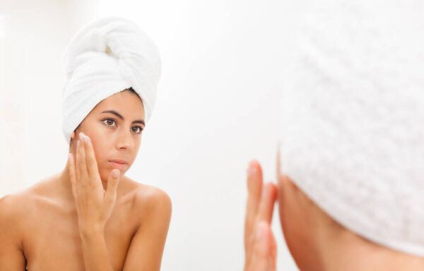 Portrait of a beautiful young woman applying face cream and looking at mirror in bathroom. Youthful girl with perfect skin using facial cosmetics. Skin care and health concept